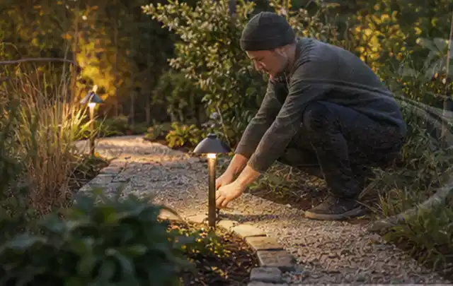 Person richtet eine niedrige warm leuchtende Gartenlampe am Kiesweg aus; Thema Smoker, Schwerpunkt Reparatur; Umsetzung.