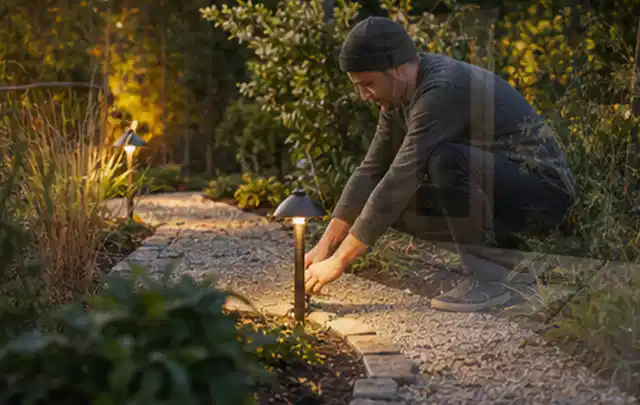 Person richtet eine niedrige warm leuchtende Gartenlampe am Kiesweg aus; Thema Smoker, Schwerpunkt Sicherheit; Umsetzung.