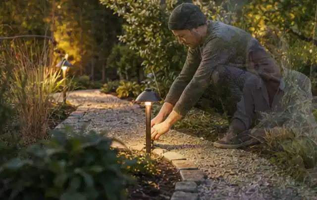 Person richtet eine niedrige warm leuchtende Gartenlampe am Kiesweg aus; Thema Sonnenschirme, Schwerpunkt Nachhaltigkeit; Kontrolle.