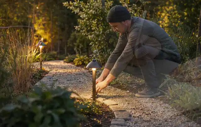 Person richtet eine niedrige warm leuchtende Gartenlampe am Kiesweg aus; Thema Sonnenschirme, Schwerpunkt Outdoor Freizeit; Ergebnis.