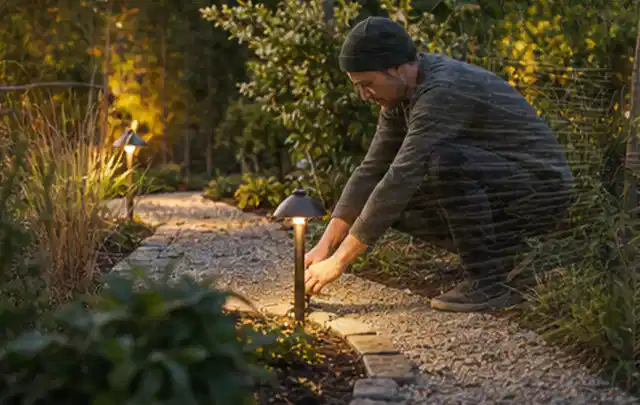 Person richtet eine niedrige warm leuchtende Gartenlampe am Kiesweg aus; Thema Sonnenschirme, Schwerpunkt Reinigung; Umsetzung.
