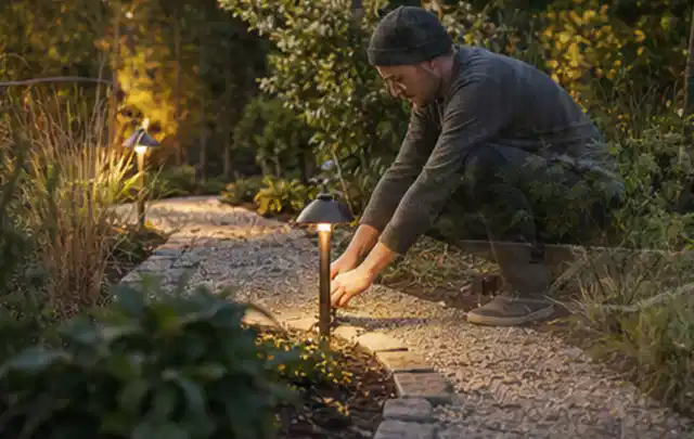 Person richtet eine niedrige warm leuchtende Gartenlampe am Kiesweg aus; Thema Sonnenschirme, Schwerpunkt Winterlager; Kontrolle.