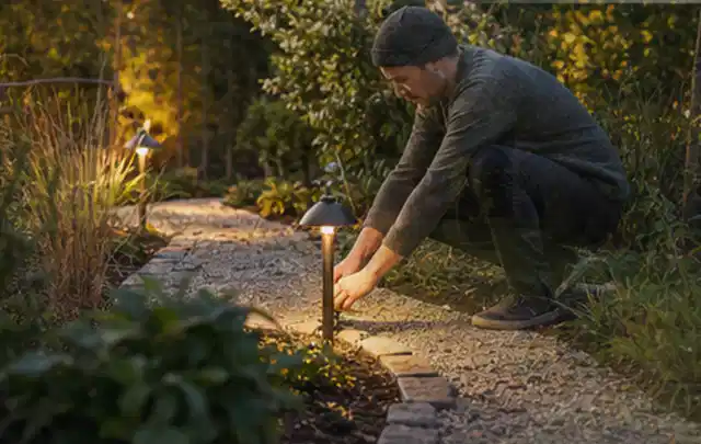 Person richtet eine niedrige warm leuchtende Gartenlampe am Kiesweg aus; Thema Steingarten, Schwerpunkt Materialwahl; Ausgangslage.