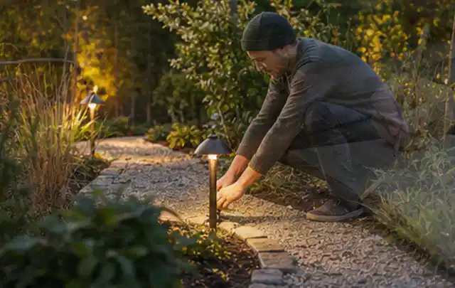 Person richtet eine niedrige warm leuchtende Gartenlampe am Kiesweg aus; Thema Steingarten, Schwerpunkt Naturgarten; Ausgangslage.