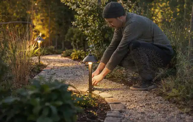 Person richtet eine niedrige warm leuchtende Gartenlampe am Kiesweg aus; Thema Steingarten, Schwerpunkt Pflegeleichtigkeit; Praxisalltag.