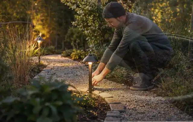 Person richtet eine niedrige warm leuchtende Gartenlampe am Kiesweg aus; Thema Steingarten, Schwerpunkt Privatsphäre; Umsetzung.