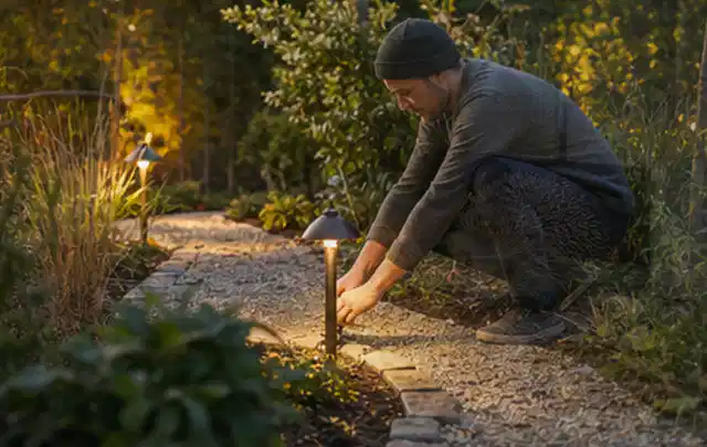 Person richtet eine niedrige warm leuchtende Gartenlampe am Kiesweg aus; Thema Steingarten, Schwerpunkt Wegeführung; Umsetzung.