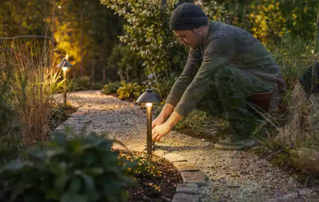 Person richtet eine niedrige warm leuchtende Gartenlampe am Kiesweg aus; Thema Stihl iMow, Schwerpunkt Ersatzteile; Umsetzung.