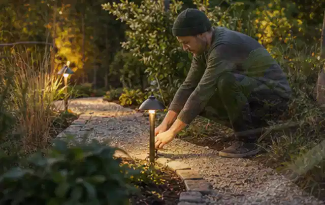 Person richtet eine niedrige warm leuchtende Gartenlampe am Kiesweg aus; Thema Stihl iMow, Schwerpunkt Fehlerhilfe; Praxisalltag.