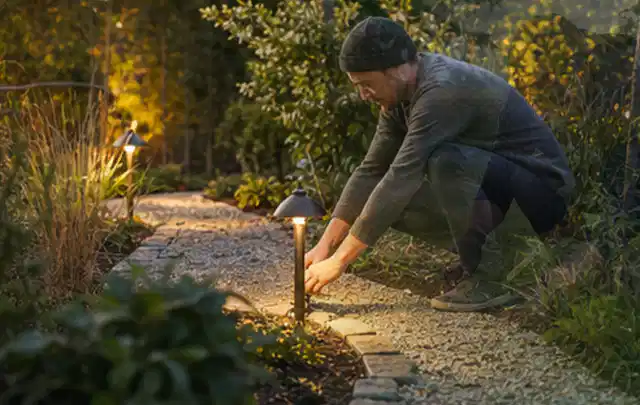 Person richtet eine niedrige warm leuchtende Gartenlampe am Kiesweg aus; Thema Stihl iMow, Schwerpunkt Mähroboter; Ausgangslage.