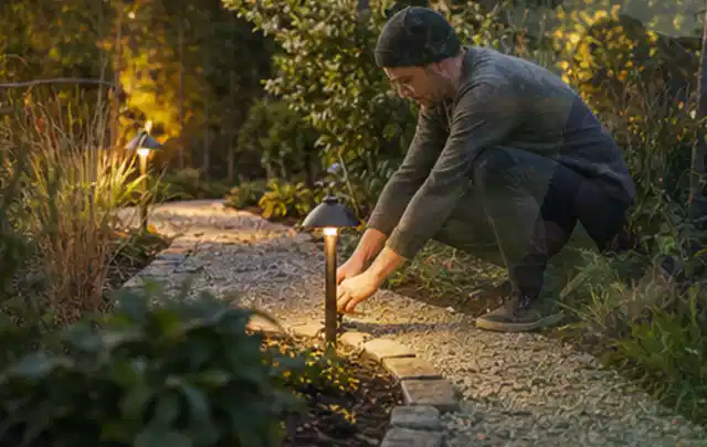 Person richtet eine niedrige warm leuchtende Gartenlampe am Kiesweg aus; Thema Stihl iMow, Schwerpunkt Reparatur; Ausgangslage.