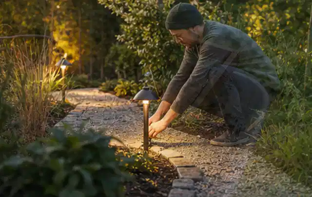 Person richtet eine niedrige warm leuchtende Gartenlampe am Kiesweg aus; Thema Terrasse, Schwerpunkt Anleitung; Ausgangslage.