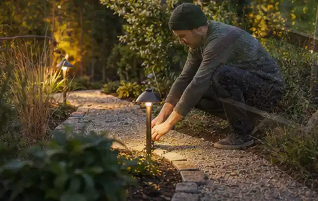 Person richtet eine niedrige warm leuchtende Gartenlampe am Kiesweg aus; Thema Terrasse, Schwerpunkt Bepflanzung; Praxisalltag.