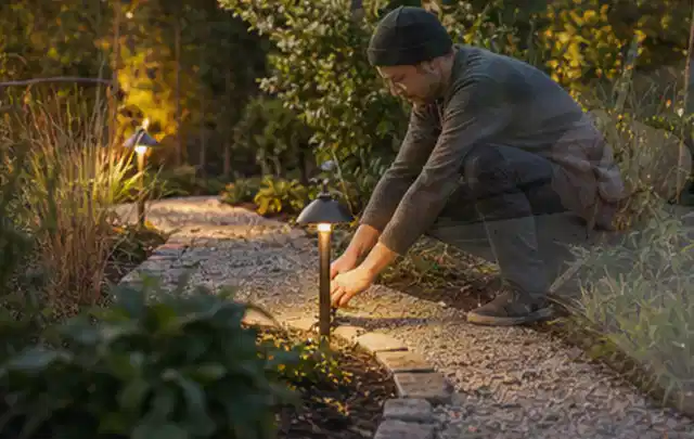 Person richtet eine niedrige warm leuchtende Gartenlampe am Kiesweg aus; Thema Terrasse, Schwerpunkt Fehlerhilfe; Ausgangslage.