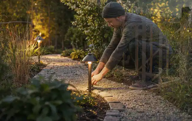 Person richtet eine niedrige warm leuchtende Gartenlampe am Kiesweg aus; Thema Terrasse, Schwerpunkt Materialwahl; Praxisalltag.