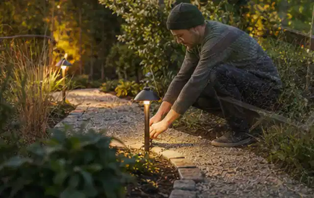 Person richtet eine niedrige warm leuchtende Gartenlampe am Kiesweg aus; Thema Terrasse, Schwerpunkt Naturgarten; Ausgangslage.