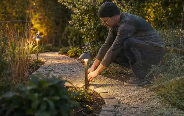 Person richtet eine niedrige warm leuchtende Gartenlampe am Kiesweg aus; Thema Terrasse, Schwerpunkt Wegeführung; Ergebnis.
