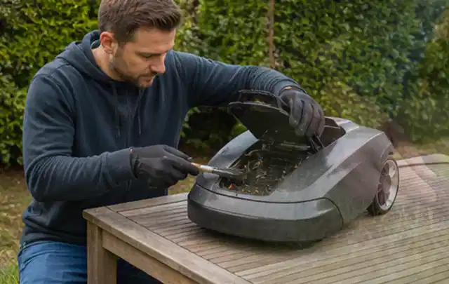 Person wartet einen geöffneten Mähroboter auf einem stabilen Gartentisch; Thema Vertikutierer, Schwerpunkt Ersatzteile; Ausgangslage.