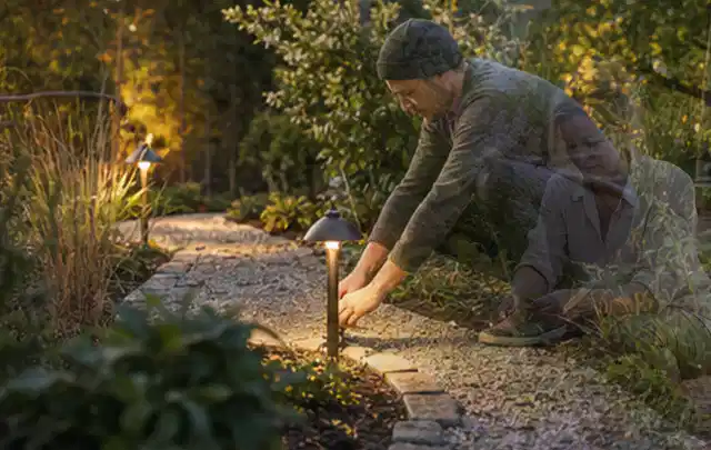 Person richtet eine niedrige warm leuchtende Gartenlampe am Kiesweg aus; Thema Vorgarten, Schwerpunkt Bepflanzung; Praxisalltag.