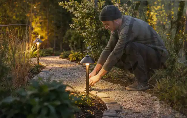 Person richtet eine niedrige warm leuchtende Gartenlampe am Kiesweg aus; Thema Vorgarten, Schwerpunkt Checkliste; Praxisalltag.