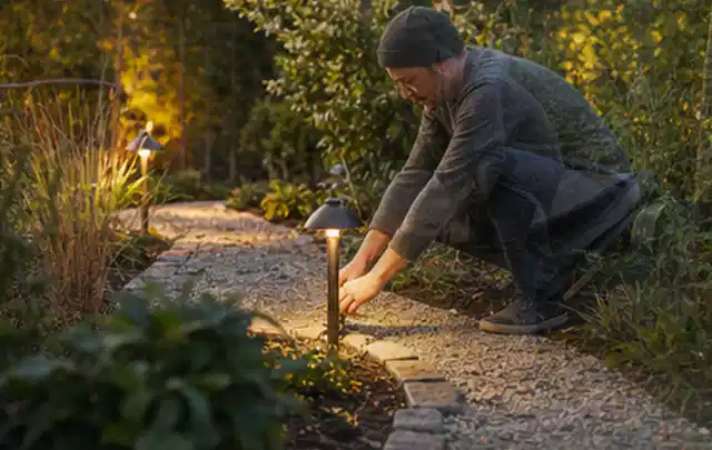 Person richtet eine niedrige warm leuchtende Gartenlampe am Kiesweg aus; Thema Vorgarten, Schwerpunkt Gärten mit kleiner Fläche; Praxisalltag.