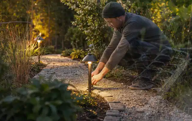 Person richtet eine niedrige warm leuchtende Gartenlampe am Kiesweg aus; Thema Vorgarten, Schwerpunkt Garten gestalten; Kontrolle.