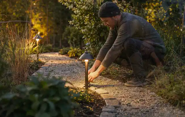 Person richtet eine niedrige warm leuchtende Gartenlampe am Kiesweg aus; Thema Vorgarten, Schwerpunkt Inspiration; Ausgangslage.