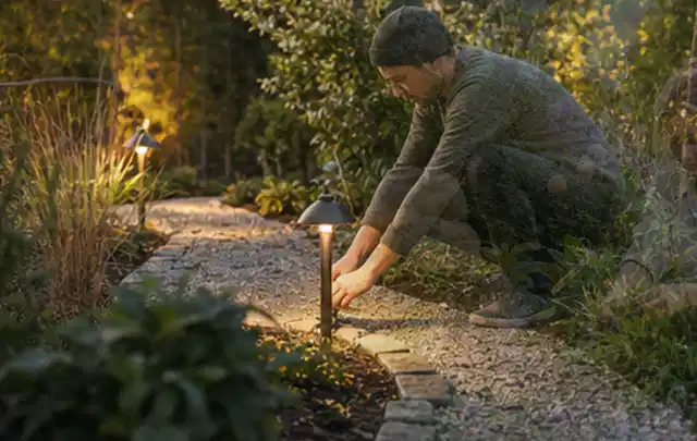 Person richtet eine niedrige warm leuchtende Gartenlampe am Kiesweg aus; Thema Vorgarten, Schwerpunkt Naturgarten; Umsetzung.