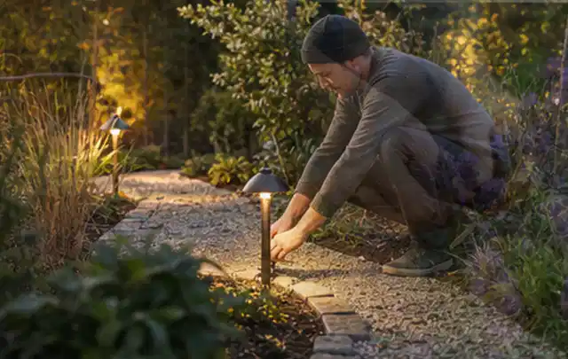 Person richtet eine niedrige warm leuchtende Gartenlampe am Kiesweg aus; Thema Vorgarten, Schwerpunkt Pflegeleichtigkeit; Umsetzung.