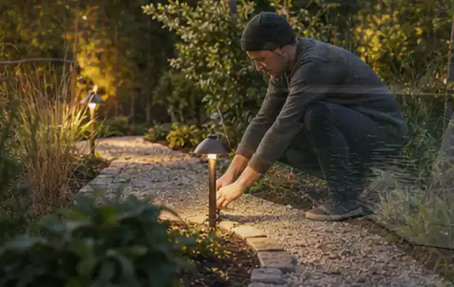 Person richtet eine niedrige warm leuchtende Gartenlampe am Kiesweg aus; Thema Wege, Schwerpunkt Garten gestalten; Umsetzung.