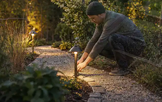 Person richtet eine niedrige warm leuchtende Gartenlampe am Kiesweg aus; Thema Whirlpools, Schwerpunkt Dimensionierung; Kontrolle.