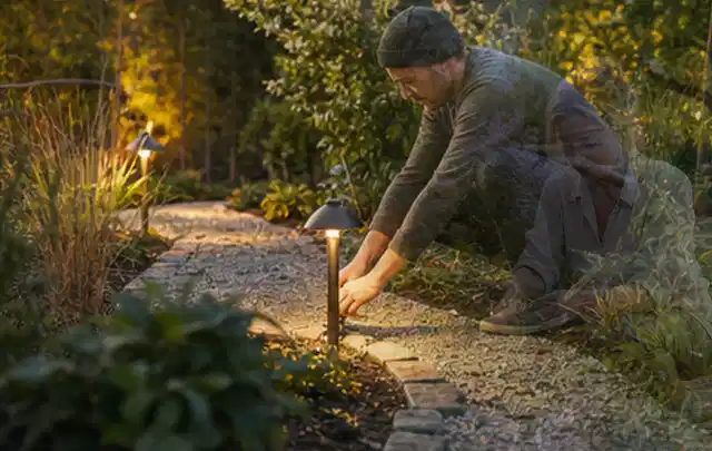 Person richtet eine niedrige warm leuchtende Gartenlampe am Kiesweg aus; Thema Whirlpools, Schwerpunkt Gärten mit kleiner Fläche; Praxisalltag.