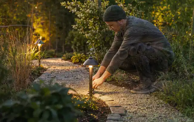 Person richtet eine niedrige warm leuchtende Gartenlampe am Kiesweg aus; Thema Whirlpools, Schwerpunkt Reparatur; Umsetzung.