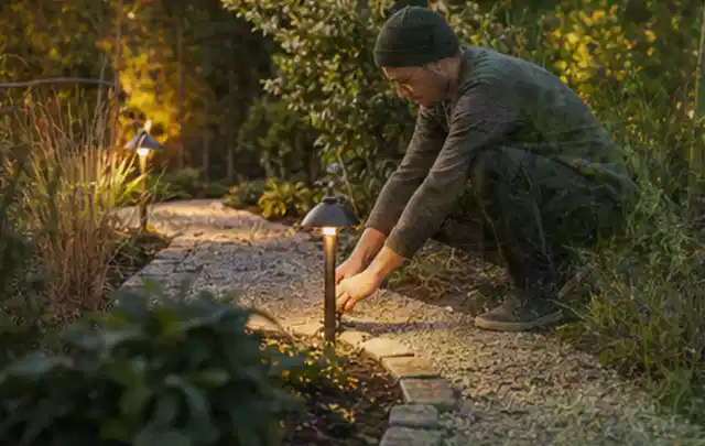 Person richtet eine niedrige warm leuchtende Gartenlampe am Kiesweg aus; Thema Whirlpools, Schwerpunkt Sicherheit; Ausgangslage.