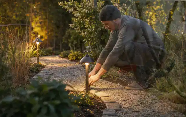 Person richtet eine niedrige warm leuchtende Gartenlampe am Kiesweg aus; Thema Whirlpools, Schwerpunkt Systemvergleich; Ergebnis.