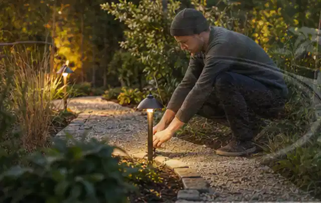 Person richtet eine niedrige warm leuchtende Gartenlampe am Kiesweg aus; Thema Whirlpools, Schwerpunkt Testkriterien; Praxisalltag.