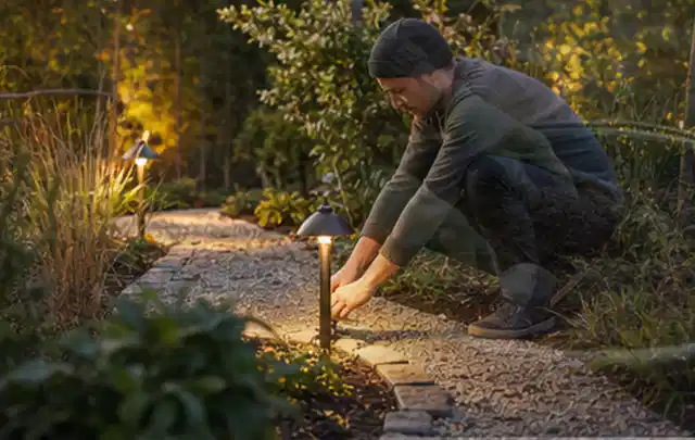 Person richtet eine niedrige warm leuchtende Gartenlampe am Kiesweg aus; Thema Whirlpools, Schwerpunkt Vergleich; Kontrolle.