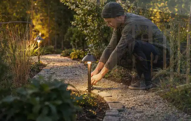 Person richtet eine niedrige warm leuchtende Gartenlampe am Kiesweg aus; Thema Whirlpools, Schwerpunkt Winterlager; Umsetzung.
