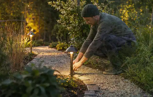 Person richtet eine niedrige warm leuchtende Gartenlampe am Kiesweg aus; Thema Worx Landroid, Schwerpunkt Checkliste; Umsetzung.