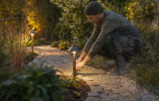 Person richtet eine niedrige warm leuchtende Gartenlampe am Kiesweg aus; Thema Worx Landroid, Schwerpunkt Gärten mit kleiner Fläche; Ausgangslage.
