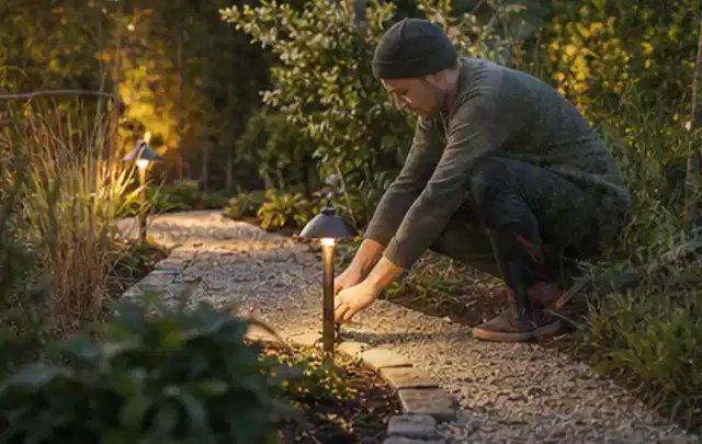 Person richtet eine niedrige warm leuchtende Gartenlampe am Kiesweg aus; Thema Worx Landroid, Schwerpunkt Sicherheit; Ausgangslage.