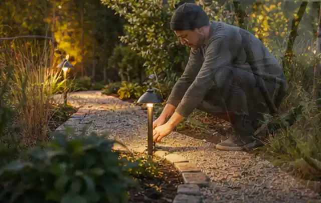 Person richtet eine niedrige warm leuchtende Gartenlampe am Kiesweg aus; Thema Worx Landroid, Schwerpunkt Systemvergleich; Ausgangslage.