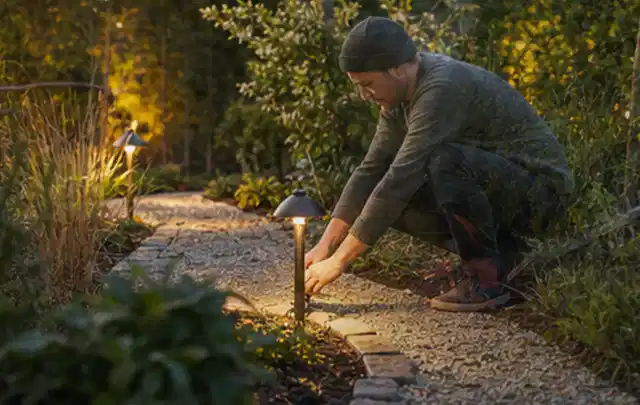 Person richtet eine niedrige warm leuchtende Gartenlampe am Kiesweg aus; Thema Worx Landroid, Schwerpunkt Testkriterien; Umsetzung.