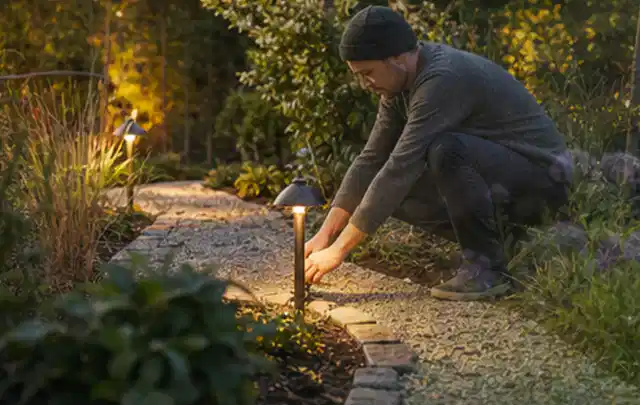 Person richtet eine niedrige warm leuchtende Gartenlampe am Kiesweg aus; Thema Worx Landroid, Schwerpunkt Winterlager; Kontrolle.