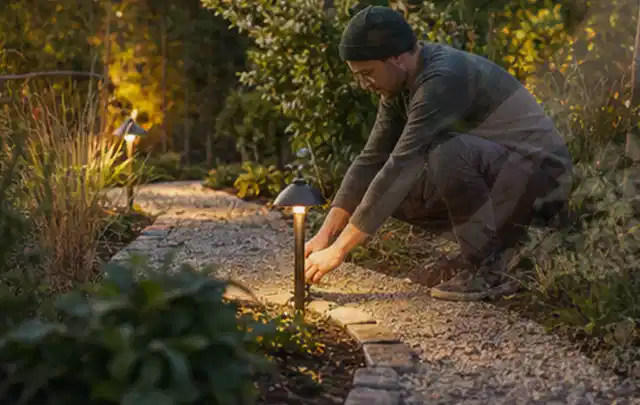 Person richtet eine niedrige warm leuchtende Gartenlampe am Kiesweg aus; Thema WPC Sichtschutz, Schwerpunkt DIY; Praxisalltag.