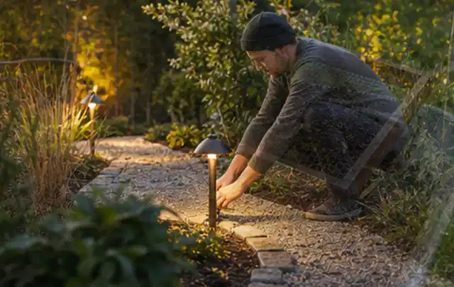 Person richtet eine niedrige warm leuchtende Gartenlampe am Kiesweg aus; Thema WPC Sichtschutz, Schwerpunkt Kosten; Ausgangslage.