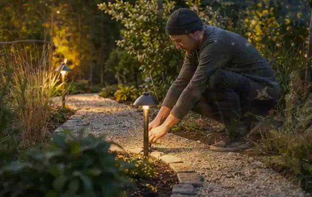 Person richtet eine niedrige warm leuchtende Gartenlampe am Kiesweg aus; Thema WPC Sichtschutz, Schwerpunkt Pflege; Kontrolle.